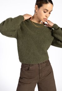 Olijfgroen, fluffy trui met een ronde hals en poffende mouwen, gecombineerd met een bruine chino. Heeft een gouden ketting als sierraad.