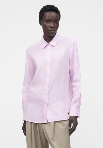 Γυναίκα που φοράει ένα ανοιχτό ροζ πουκάμισο με κουμπιά και μακριά μανίκια και μπεζ φαρδιά παντελόνια, στέκεται μπροστά σε λευκό, μονόχρωμο φόντο.