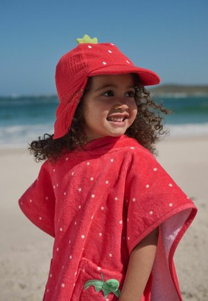 Smaidošs bērns ar cirtainiem matiem, valkājot sarkanu zemeņu tēmas cepuri un atbilstošu punktiņu pludmales pončo pie jūras saulainā dienā.