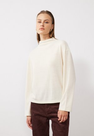 Haut crème à manches longues avec un col montant, confectionné dans un tissu doux. Associé à un pantalon en velours côtelé bordeaux foncé.