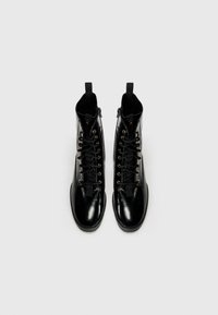 Botas altas negras con acabado brillante, con ojales dorados, cordones negros y tiradores en el collar para facilitar su uso.