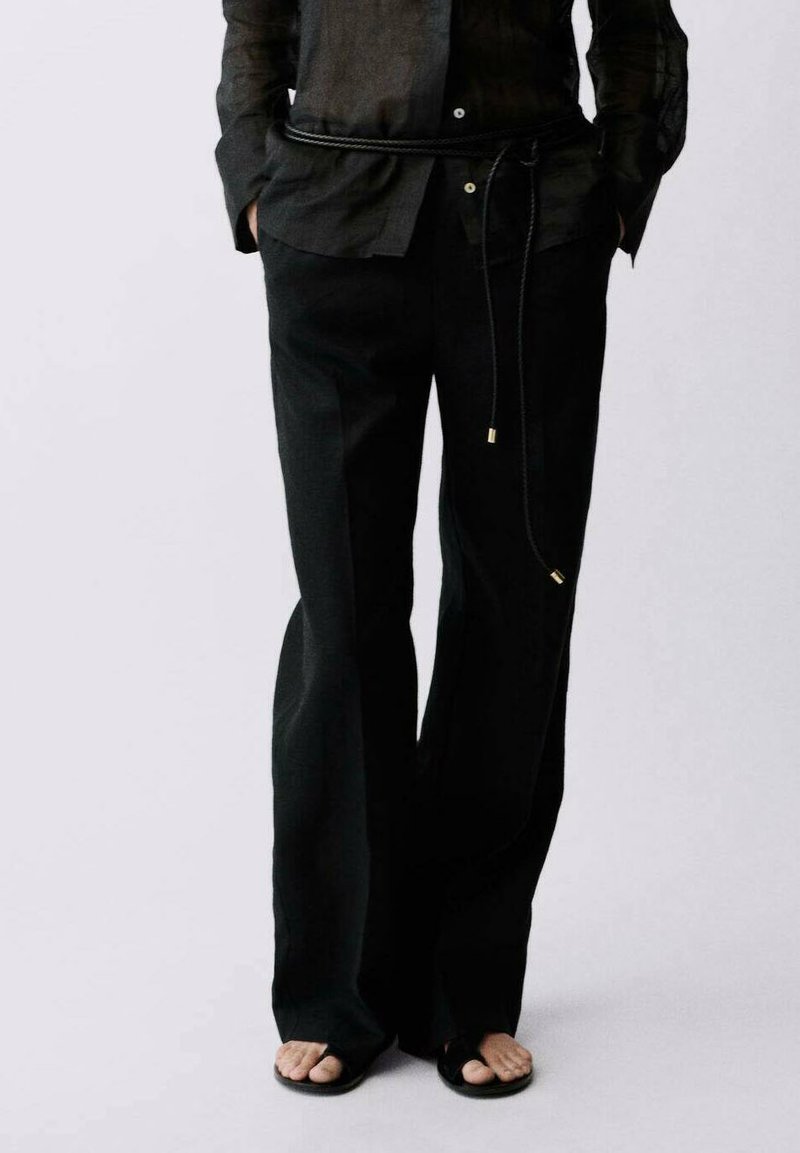 Black wide-leg pants made from a soft material, featuring a drawstring waist with gold accents. Paired with flat, open-toed sandals.
