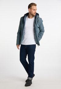 Giacca isolante blu con cappuccio, zip frontale e bottoni; indossata sopra una maglietta bianca e jeans in denim scuri con risvolti e scarpe nere.