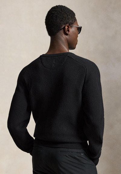 Homme portant des lunettes de soleil sombres, un pull en maille noir et un pantalon noir, debout les mains dans les poches, de dos sur un fond uni.