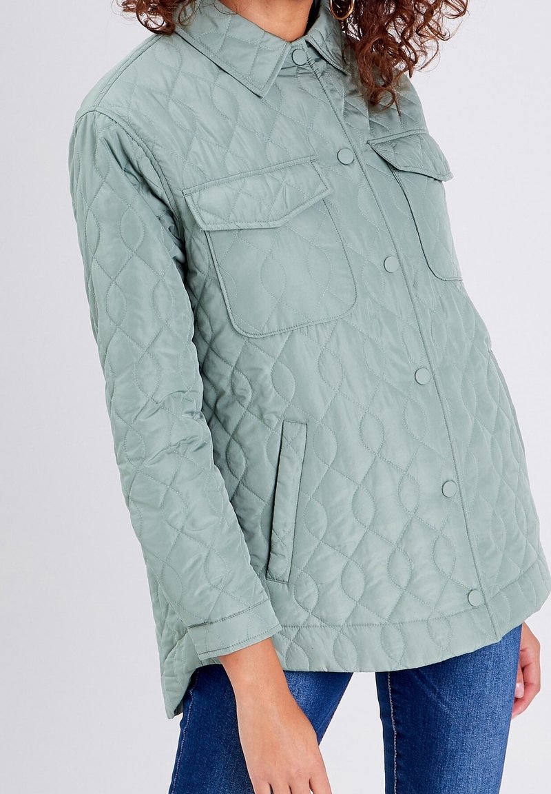 Personne portant une veste matelassée vert clair avec boutons pression, deux poches à rabat sur la poitrine et poches latérales fendues, associée à un jean bleu.