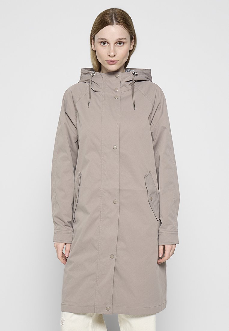 Jeune femme portant un long imperméable taupe à capuche avec des boutons-pression sur le devant et des poches à rabat sur les côtés, se tenant contre un fond uni.