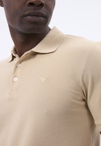 Beige Polohemd aus texturiertem Stoff mit klassischem Kragen, drei Knöpfen und einem gestickten Logo auf der linken Brust.