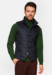 Granatowa, ocieplana kamizelka o pikowanej teksturze, z wysokim kołnierzem, z przodu zapinana na guziki oraz z bocznymi kieszeniami, noszona na zielonym swetrze.