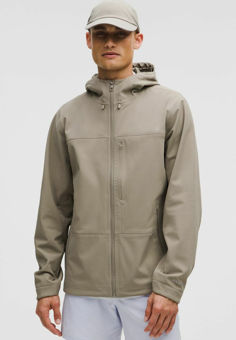 Chaqueta con capucha de un beige claro y textura suave y flexible. Cuenta con cierre de cremallera, dos bolsillos frontales y cordones ajustables en la capucha.