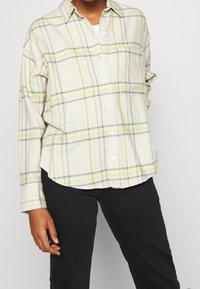 Person wearing a cream plaid button-up shirt with yellow and gray stripes and black pants against a plain light background.