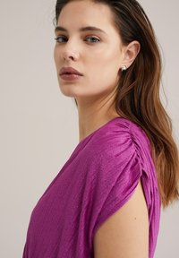 Rochie fucsia cu mâneci scurte, strânse, realizată dintr-un material texturat și strălucitor. Prim-plan al profilului modelului, evidențiind cercei minimalisti.