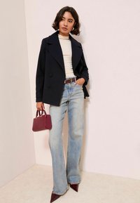 Femme portant un manteau noir croisé, un haut crème, un jean bleu clair, une ceinture bordeaux, des chaussures pointues et tenant un sac à main bordeaux.