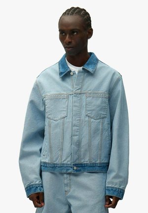 Giovane uomo con capelli intrecciati che indossa una giacca di jeans azzurro chiaro con colletto e polsini blu scuro, abbinata a pantaloni di jeans dello stesso colore.