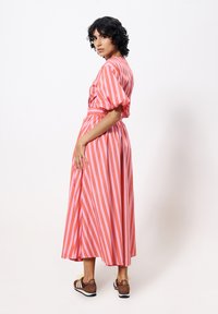 Maxikleid in Pink mit vertikalen roten Streifen, gepufften Ärmeln, gebundenem Taillenbereich und fließender Silhouette. Kombiniert mit braunen Sneakers.