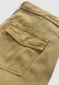 Beige denimstof med en baglomme med en syet klap og et rødt mærke. Texturen er let ribbet, med synlige syninger.