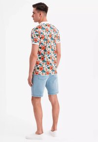 Blommig kortärmad skjorta med orange och gröna blommor på en ljus bakgrund, i kombination med ljusblå shorts och vita slip-on skor.