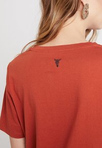 Rustfarvet t-shirt med en afslappet pasform, der har et lille sort grafisk motiv af et hoved på den øverste del af ryggen. Blødt bomuldsstof, enkelt design.