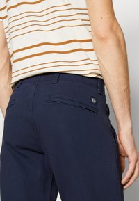 Marinblå bomullbyxor med slim fit, med en bakficka och sydda detaljer. Bärs med en skjorta med kräm- och brunrand.