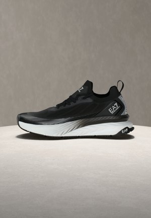 Black and gray EA7 sneaker with thick white sole, lace-up front, and pull tab on heel on a beige surface with plain background.