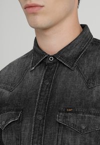 Svart denimskjorta med krage, bröstfickor, tryckknappar och ett subtilt strukturerat mönster. M ärkslabel synlig på fickan.