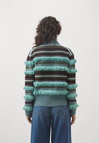 Neulottu neulepaita, jossa on vaakasuorat raidat väriltään sinivihreä, burgundi ja ruskea, korostettuna tekstuurisilla hapsuilla sekä joustinneuloksella olevat helmassa ja hihansuissa.