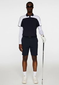 Herrarnas golfklädsel: marinblå pikétröja med kort ärm och vita detaljer, marinblå shorts, vita strumpor och vita skor. Håller i en golfklubba och bär solglasögon.