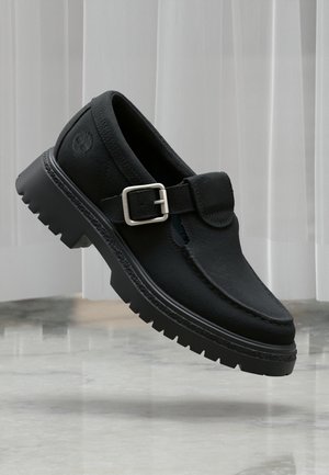 Scarpa con fibbia in pelle nera con suola spessa scolpita, cuciture moc-toe e logo in rilievo sul lato del tallone, mostrata sopra un pavimento in marmo.