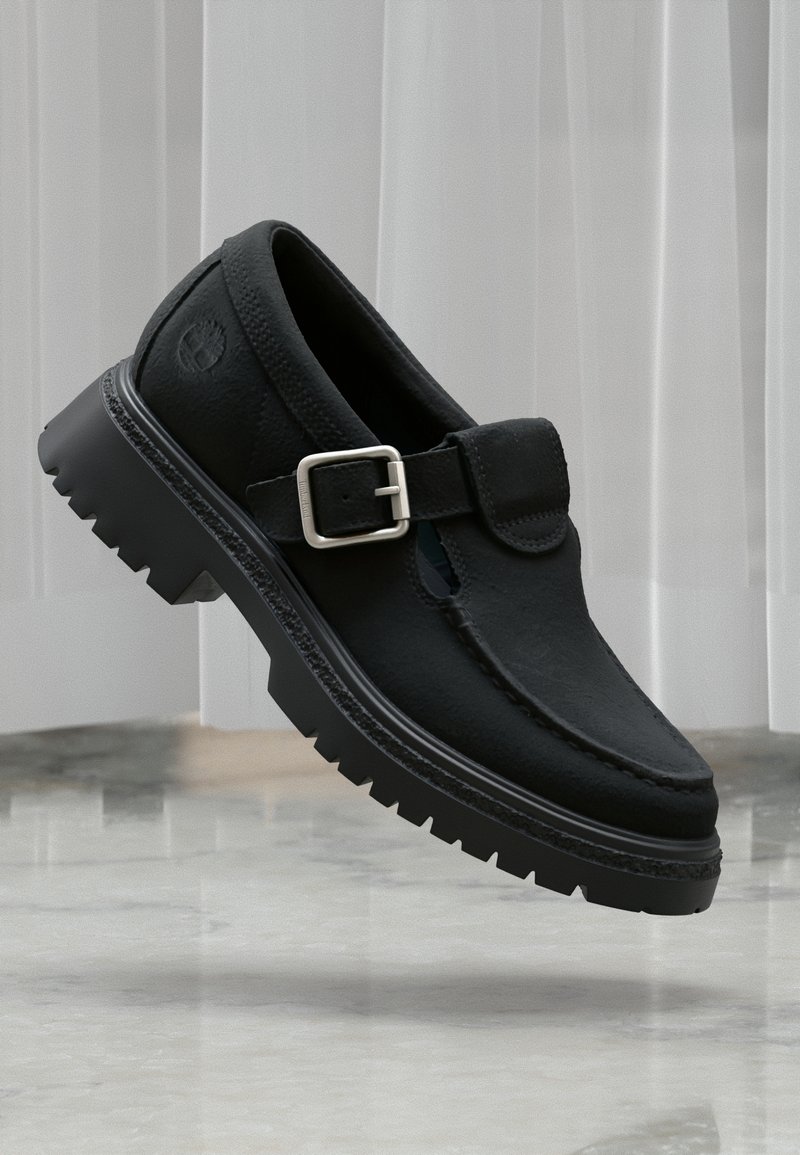 Μαύρο δερμάτινο παπούτσι με αγκράφα, χοντρή σόλα με αυλακώσεις, ραφή moc-toe και ανάγλυφο λογότυπο στο πλευρό της φτέρνας, τοποθετημένο πάνω σε μαρμάρινο πάτωμα.