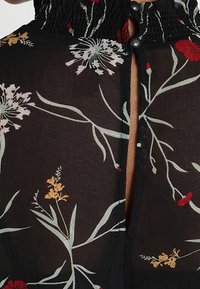 Czarna bluzka w kwiaty z marszczonym dekoltem i zapięciem na guziki. Posiada kolorowe wzory kwiatowe w odcieniach czerwieni, żółci i zieleni na przezroczystym materiale.