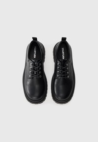 Paire de chaussures à lacets en cuir noir avec semelles épaisses, vues de dessus, sur un fond gris clair.