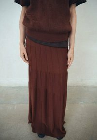 Personne portant un haut en tricot côtelé marron et une jupe longue assortie, se tenant sur un sol en béton contre un mur gris uni.