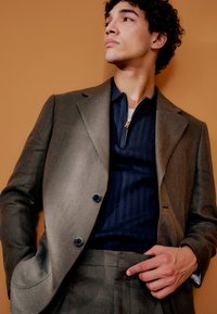 Fiatal férfi göndör hajjal, barna öltönyben és tengerészkék cipzáras pulóverben, egy egyszínű narancssárga háttér előtt pózol.