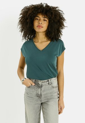 Femme aux cheveux bouclés portant un haut bleu sarcelle à col en V, un jean gris clair et des bracelets argentés, debout avec une main dans la poche.