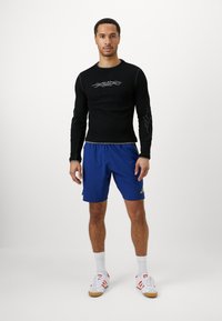 Camisola atlética preta de mangas longas com um design tribal, emparelhada com calções azuis que apresentam um logótipo amarelo. O modelo usa meias e sapatilhas brancas.