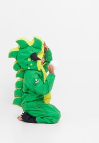 Groene dinosaurus-thema jas met gele accenten, gestructureerde stekels en zwarte details. Kind zit, houdt een witte ronde object vast.