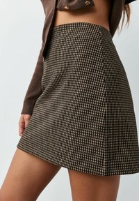 Brun og beige mininederdel med hundetandsmønster, båret med en brun kort cardigan, der viser maven og armen.