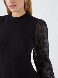 Γυναίκα που φοράει μια μαύρη ριμπ μπλούζα με ψηλό λαιμό και διαφανή φουσκωτά μανίκια, κεντημένα με μαύρα floral σχέδια.