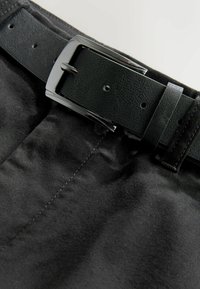 Fekete bőrbelt fényes ezüst csattal, szorosan illeszkedve fekete szövetnadrághoz, kiemelve a sima textúrát és a letisztult vonalakat.