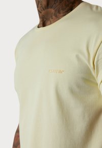 T-shirt jaune clair en coton doux, avec un col rond, des manches courtes et un logo en relief discret de la même couleur.