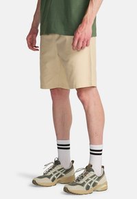 Mężczyzna ubraný w beżowe krótkie spodenki do kolan, białe skarpetki z czarnymi paskami oraz szare i beżowe buty sportowe, stojący na jednolitym tle.