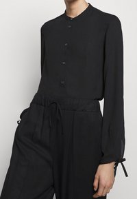 Blouse noire à manches longues avec un col rond et des fermetures à boutons, tissu semblant léger. Associée à un pantalon noir à cordon de serrage.