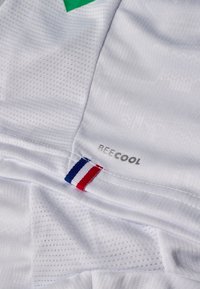 Tessuto bianco traspirante con la scritta "BEE COOL" e una piccola etichetta cucita con strisce verticali blu, bianche e rosse.