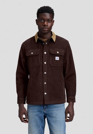 Homme portant une veste en velours côtelé marron avec des poches avant et un col beige, associée à un jean bleu clair, debout devant un fond gris.