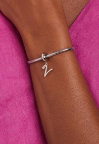 Bracelet en argent avec un design de chaîne serpent, présentant un petit charme en forme de lettre suspendu et un accent en pierre précieuse transparente. Fond en tissu rose.