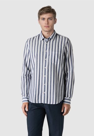 Junger Mann, der ein langärmliges weißes Hemd mit vertikalen grauen Streifen und dunkle Hose trägt, vor einem schlichten hellgrauen Hintergrund steht.