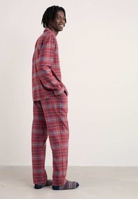 Hymyilevä mies seisoo kädet taskuissa punaisen ruudullisen pyjaman ja värikkäiden kuvioiden sukkien kanssa yksinkertaista vaaleaa taustaa vasten.