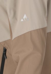 Lightweight, waterproof jacket featuring a tan body and beige upper section. Includes a silver logo and a zippered pocket on the side.