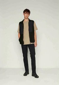 Must must vest must, must beige t-särk, must püksid ja mustad jalanõud. Vestil on tekstureeritud disain. Lihtne taust.