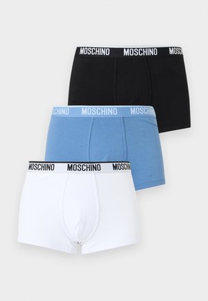 Hemdloze man staat op blote voeten, draagt blauwe Moschino boxershorts tegen een effen witte achtergrond.