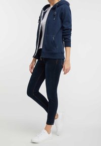 Felpa con zip blu navy dotata di coulisse, tasche frontali, indossata sopra una camicia bianca, abbinata a jeans skinny scuri e scarpe da ginnastica bianche.
